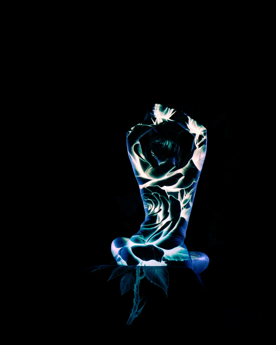 A seated person with arms raised above the head, illuminated by a blue and white floral pattern projected onto their back and surrounding dark space.